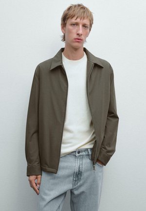 Junger Mann in einer olivgrünen Zip-Jacke über einem weißen Hemd und hellblauen Jeans, der vor einem schlichten hellen Hintergrund steht.