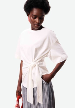 Femme aux cheveux courts et bouclés portant un haut blanc à manches courtes noué, un pantalon rayé, des boucles d'oreilles rouges, et tenant un sac rouge à motifs.