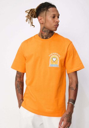 Homme avec des dreadlocks et des tatouages portant un t-shirt orange avec un motif de cœur et de vague marqué "Farniente", debout devant un fond blanc.