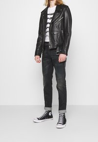 Chaqueta de motorista de cuero negro con cremalleras plateadas, superpuesta sobre una camiseta gráfica blanca. Usada con jeans negros descoloridos y zapatillas altas negras.