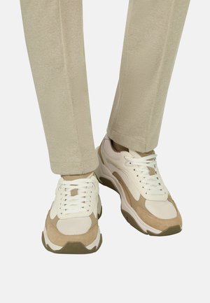 Beige en witte sneakers met dikke zolen, gedragen met lichtbeige broek, frontaal weergegeven op mid-kuitniveau tegen een effen achtergrond.
