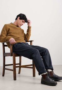 Brauner Strickpullover mit geripptem Muster, dunkelblaue Hose und braune Ankle Boots mit elastischen Akzenten, sitzend auf einem Holzstuhl.