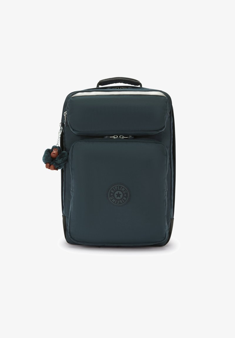 Donkerblauwe stof rugzak met bovenhandvat, voorzak met rits, logo-patch en een kleine harige aap sleutelhanger eraan bevestigd.