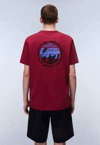 Burgunderfarbendes Baumwoll-T-Shirt mit einem runden Grafikdesign auf der Rückseite, das Bäume und Berge in Blau und Schwarz zeigt, mit dem Text "Napapijri Geographic".