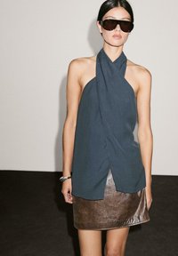 Donna che indossa occhiali da sole oversize scuri, top halter senza maniche scuro, mini gonna metallica marrone e bracciale argentato, in piedi contro uno sfondo neutro.