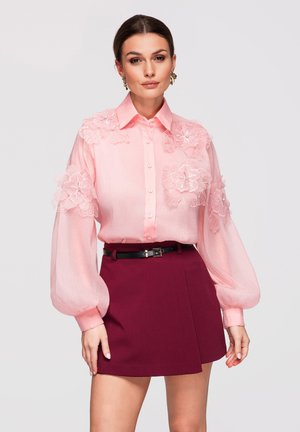 Kobieta w przezroczystej różowej bluzce z kwiatowymi aplikacjami i bufkami na rękawach, w połączeniu z wysokim stanem bordowej spódnicy i czarnym paskiem.