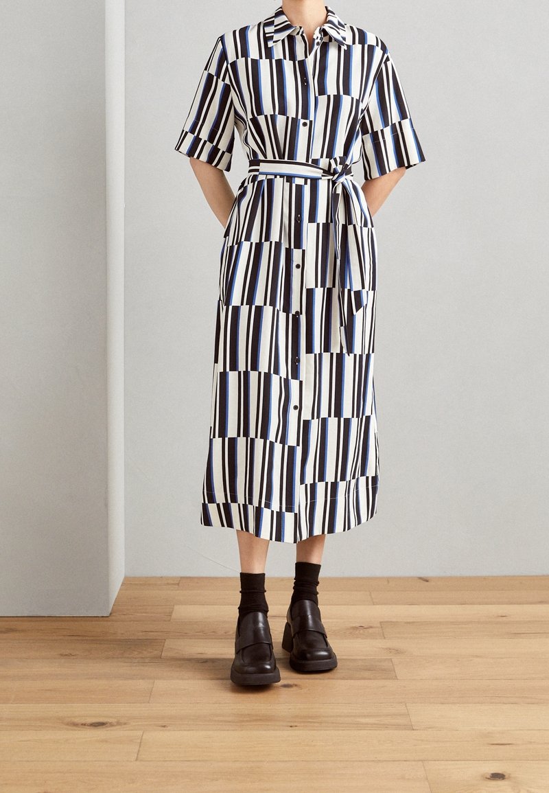 Abito camicia a righe in nero, bianco e blu con maniche corte, apertura con bottoni e vita cinte. Presenta una tasca a toppa e un taglio a metà gamba.