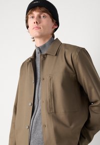 Brun knappjakke med krage, enkelt brystlomme, kombinert med en grå zip-up genser. Stoffet ser glatt og strukturert ut.