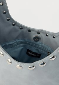 Sac en similicuir bleu orné de clous métalliques autour de l'ouverture, doté d'un intérieur en tissu doux et d'une poche zippée. Étiquette visible à l'intérieur.