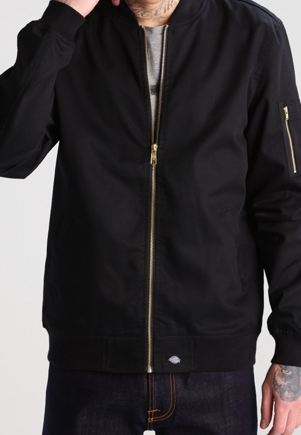 Homme portant une veste bomber noire zippée avec des fermetures éclair dorées et un jean bleu foncé, main levée près de la tête.