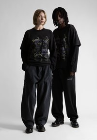 Két modell áll egymás mellett fekete, rétegezett koponya grafikás pólókban, laza fekete nadrágban és fekete cipőben, világos, egyszínű háttér előtt.