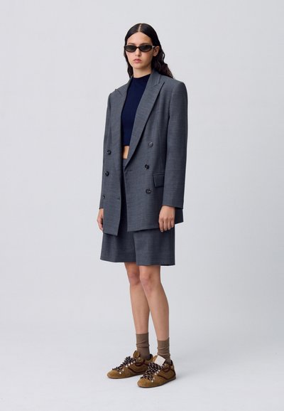 Blazer grigio doppio petto con bottoni grandi, abbinato a un top corto blu navy e pantaloni corti grigi su misura. Sneakers marroni con lacci a fantasia.