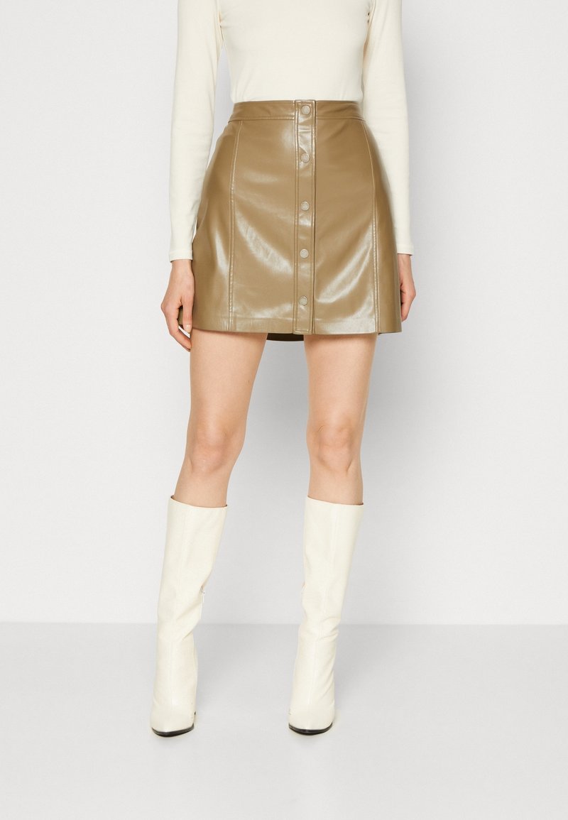 Femme portant une mini-jupe en cuir marron brillant boutonnée, un haut blanc à manches longues et des bottes blanches montantes jusqu'aux genoux, debout sur un fond blanc.