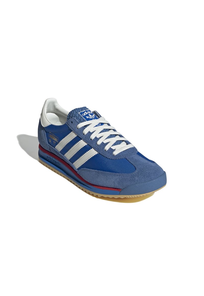 Baskets de sport bleues en suède avec des rayures blanches et des lacets, comportant une ligne d'accent rouge et une semelle en caoutchouc texturée. Design rétro classique.