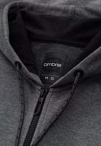 Sudadera gris con cremallera hecha de un tejido texturizado, con un interior negro, cordón y una etiqueta con el logotipo "ombre". Talla M, EUR 48.
