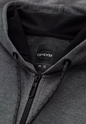 Sudadera con cremallera en tejido texturizado negro y gris. Cuenta con capucha con cordones y una etiqueta marcada "ombre, M, EUR 48."