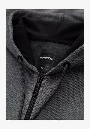 Sweat à capuche zippé en tissu texturé noir et gris. Comprend une capuche avec cordon de serrage et une étiquette indiquant "ombre, M, EUR 48".