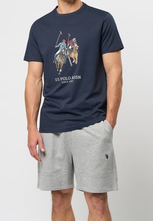Mężczyzna noszący granatowy T-shirt z dwoma zawodnikami polo na koniach oraz szare spodenki z małym wyhaftowanym logo na lewej nodze.