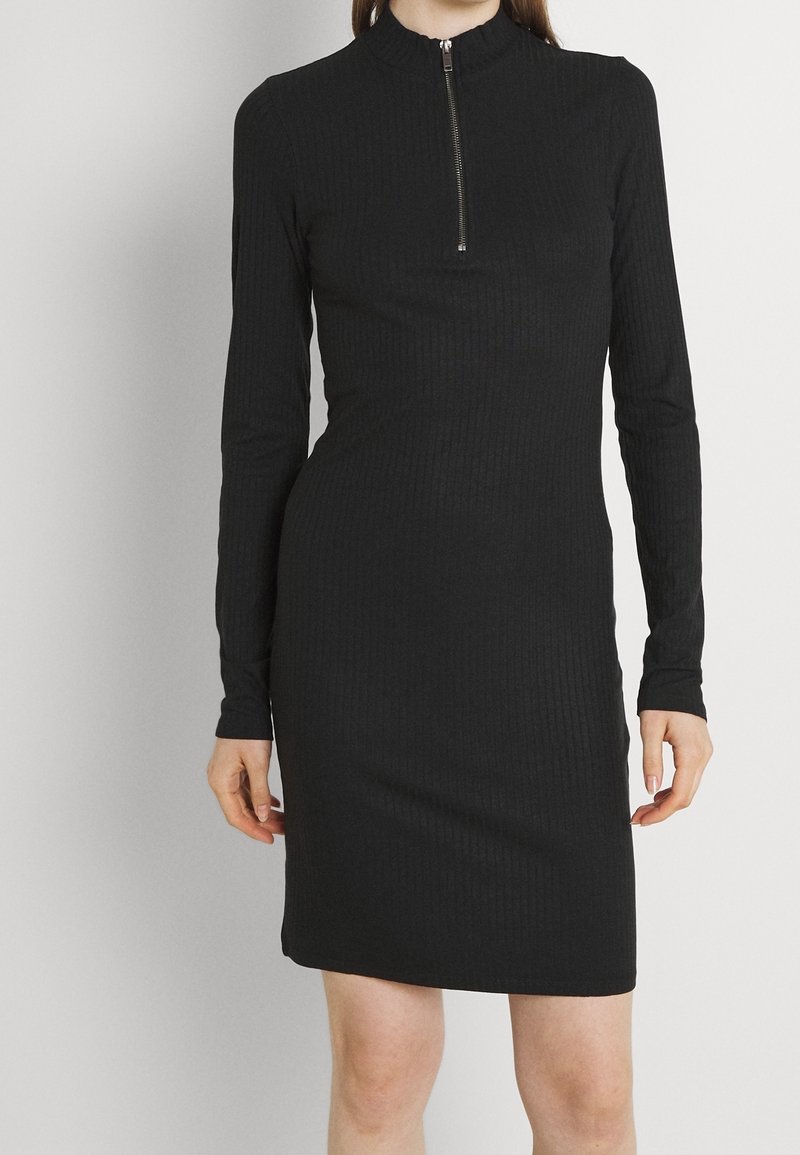 Svart ribbad klänning med långa ärmar och hög krage. Har en detalj med dragkedja framtill och en figurnära silhuett. Slät textur.