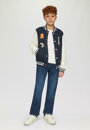 Veste varsity bleu marine et crème avec des écussons, chemise blanche, jeans bleu foncé et baskets blanches. La veste a des poignets et un col rayés.