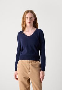 Maglione a V blu navy realizzato in tessuto morbido, con polsini e orlo a costine, e un piccolo logo ricamato sul lato sinistro del petto. Abbinato a pantaloni beige.