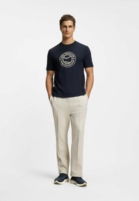 Marineblaues T-Shirt mit rundem Halsausschnitt und cremefarbenem Tennis-Logo, kombiniert mit hellbeigen, maßgeschneiderten Hosen und dunklen Sneakern.