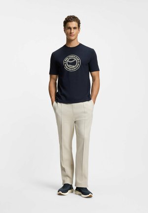 Granatowy t-shirt z okrągłym dekoltem i kremowym logo o tematyce tenisowej, zestawiony z jasnobeżowymi, dopasowanymi spodniami i ciemnymi trampkami.