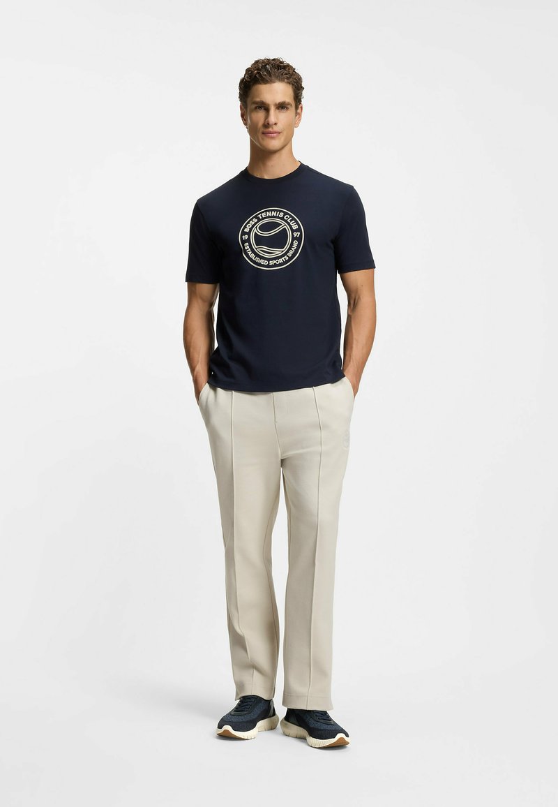 Marineblaues T-Shirt mit rundem Halsausschnitt und cremefarbenem Tennis-Logo, kombiniert mit hellbeigen, maßgeschneiderten Hosen und dunklen Sneakern.