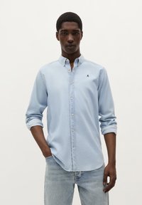 Lichtblauw overhemd met kraag, opgerolde mouwen en een klein marineblauw logo op de borst; gecombineerd met lichtdenim jeans.