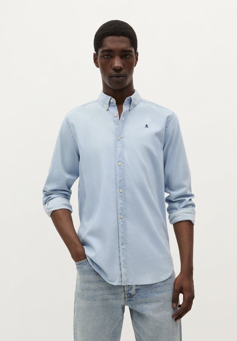 Lichtblauw overhemd met kraag, opgerolde mouwen en een klein marineblauw logo op de borst; gecombineerd met lichtdenim jeans.