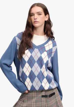 Ung kvinde med langt brunt hår iført en blå og hvid argyle sweater og ternet nederdel med sort bælte, poserer med den ene hånd på hoften.