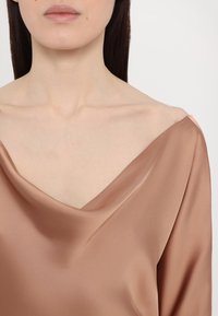 Hladká, svetlo hnedá blúzka s hlbokým výstrihom do V a splývavým golierom, ktorá ukazuje jemnú textúru a plynulý dizajn.