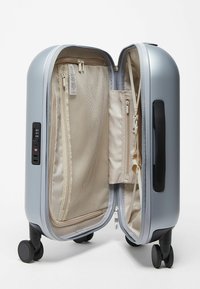Valise rigide argentée ouverte, montrant un intérieur beige avec des compartiments en filet et zippés, des sangles de compression et quatre roues noires pivotantes.