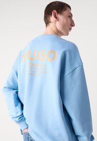 Svetlo modra kapuca iz mehkega materiala, sledi velikemu zlatemu logotipu "HUGO" na hrbtni strani, z rebrastimi manšetami in sproščenim krojem.