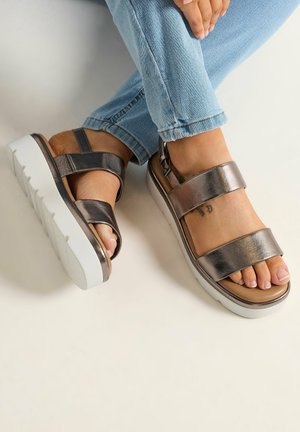 Persona che indossa sandali platform metallici con doppie fibbie, jeans azzurri e un piccolo tatuaggio visibile sul piede.