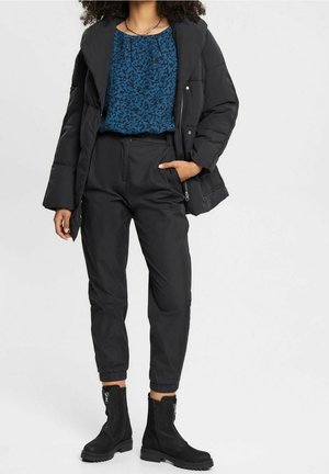 Woman wearing black puffer jacket, blue patterned blouse, black high-waist tapered pants, and black ankle boots with zippers, standing against white background.