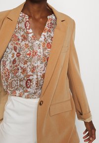Blazer beige a medida sobre una blusa con estampado floral en blanco, rojo y verde, con escote en V y detalle de botones. Se ven pantalones blancos de talle alto.