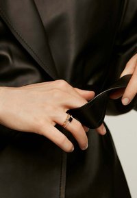 Anillo dorado y negro con una piedra negra rectangular y acentos claros, exhibido en una mano que sostiene un material negro. Prenda de cuero negro visible.