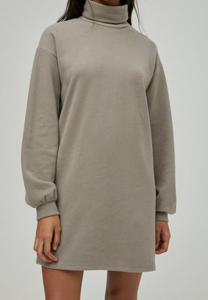 Kobieta w długim swetrze-tuniką z golfem w kolorze taupe z elastycznymi ściągaczami na rękawach, stojąca na jasnym, jednolitym tle.