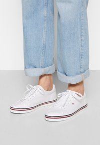Sneaker bianche in tela con striscia a contrasto blu navy e rossa sulla suola, dotate di chiusura stringata e senza hardware visibile. Indossate con jeans chiari.