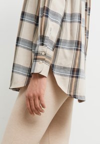 Camicia a quadri realizzata in tessuto con un motivo a scacchi beige, blu e marrone. Presenta un orlo arrotondato e polsini con bottoni.