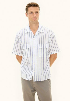 Man wearing a white short-sleeve button-up shirt with blue vertical stripes and beige pants, standing with hands behind back.