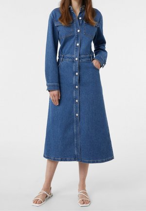 Frau trägt ein langes blaues Jeanskleid mit Knöpfen vorne, Brusttaschen und langen Ärmeln, kombiniert mit weißen, geschnürten Sandalen.