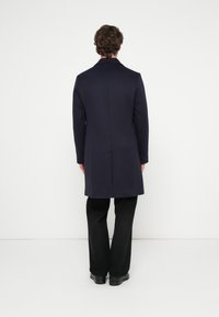 Marineblauer Mantel aus Wollmischung mit figurbetontem Schnitt, Einzelnaht am Rücken und Reverskragen, kombiniert mit schwarzen, ausgestellten Hosen und schwarzen Anzugschuhen.