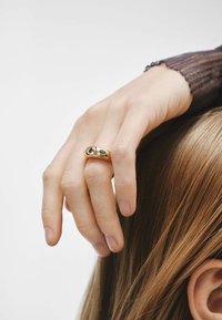 Anillo de oro con tres piedras: una piedra redonda clara, una piedra ovalada blanca y una piedra marquesa verde, llevado en una mano con pelo lacio.