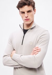 Helebeež pooltõmblukuga pullover, valmistatud pehmest kudumkangast, millel on tekstuurse kraega ja väike reljeefne logo rinnal.