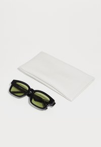 Occhiali da sole neri con lenti verdi, forma rettangolare, con una montatura spessa. Accompagnati da un astuccio protettivo bianco.