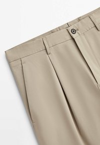 Pantalon beige avec passants de ceinture, plis à l'avant et une fermeture par un seul bouton noir sur fond blanc.