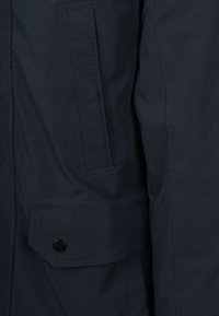 Marineblauw jack met een gestructureerde afwerking, voorzien van een klepzak die wordt gesloten met een zwarte drukknop, en contrasterende stiksel details.