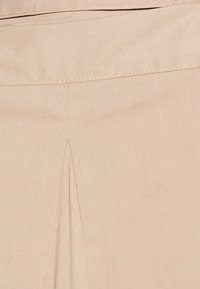Tessuto beige con una texture liscia, caratterizzato da una piega frontale e una vita dritta. Cuciture visibili lungo il bordo superiore.